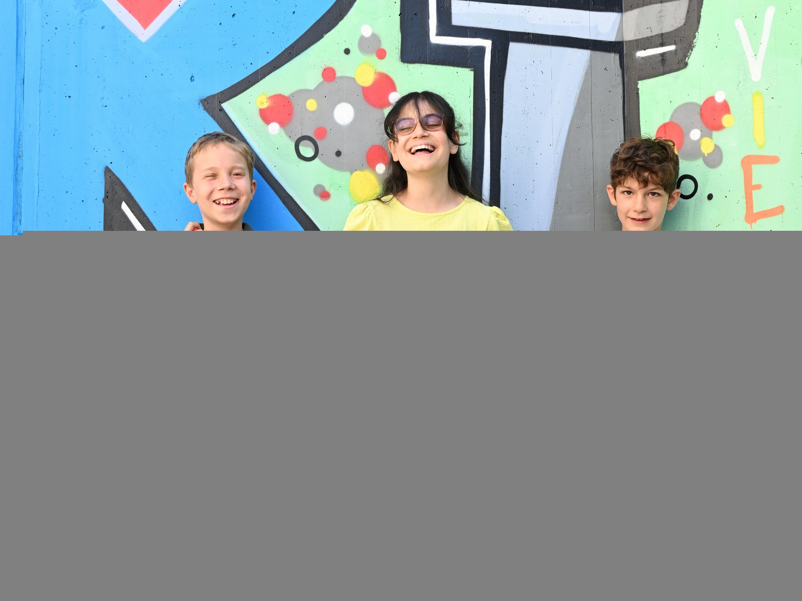 Drei Kinder stehen vor einer Wand mit einem buntem Graffiti. Ein Junge hält einen Langstock. Sie lachen.