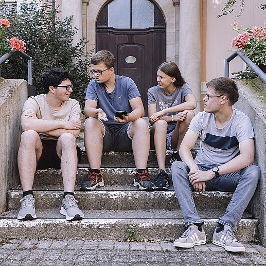 Drei Schüler und eine Schülerin sitzen draußen auf einer Treppe vor dem Eingang eines Schulgebäudes